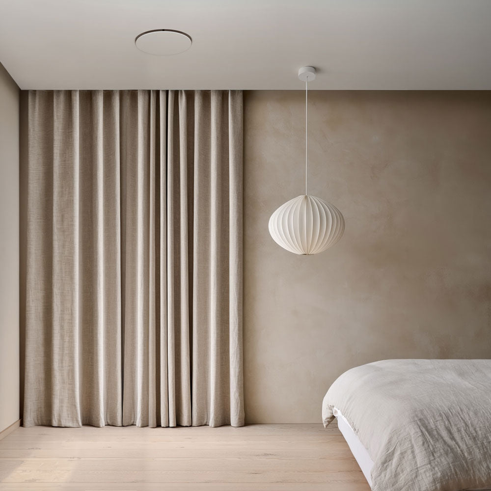 A minimalist bedroom interior with the Shadowline Diffuser AC Round installed in the ceiling, floor-length taupe linen curtains, a white ribbed pendant light, a beige plaster wall, light wood flooring, and part of a white bed visible