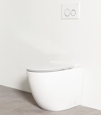 Milu Odourless Crest in-wall floor mounted toilet angled side view with white flush plate. Positioned against white tiled wall on warm grey coloured floor tiles