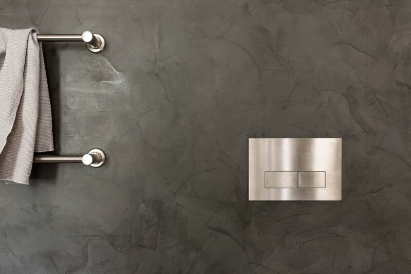 Milu Odourless Ultraslim Flush Plate in brushed stainless steel is photographed against a textured charcoal coloured plaster wall at Alexander House
